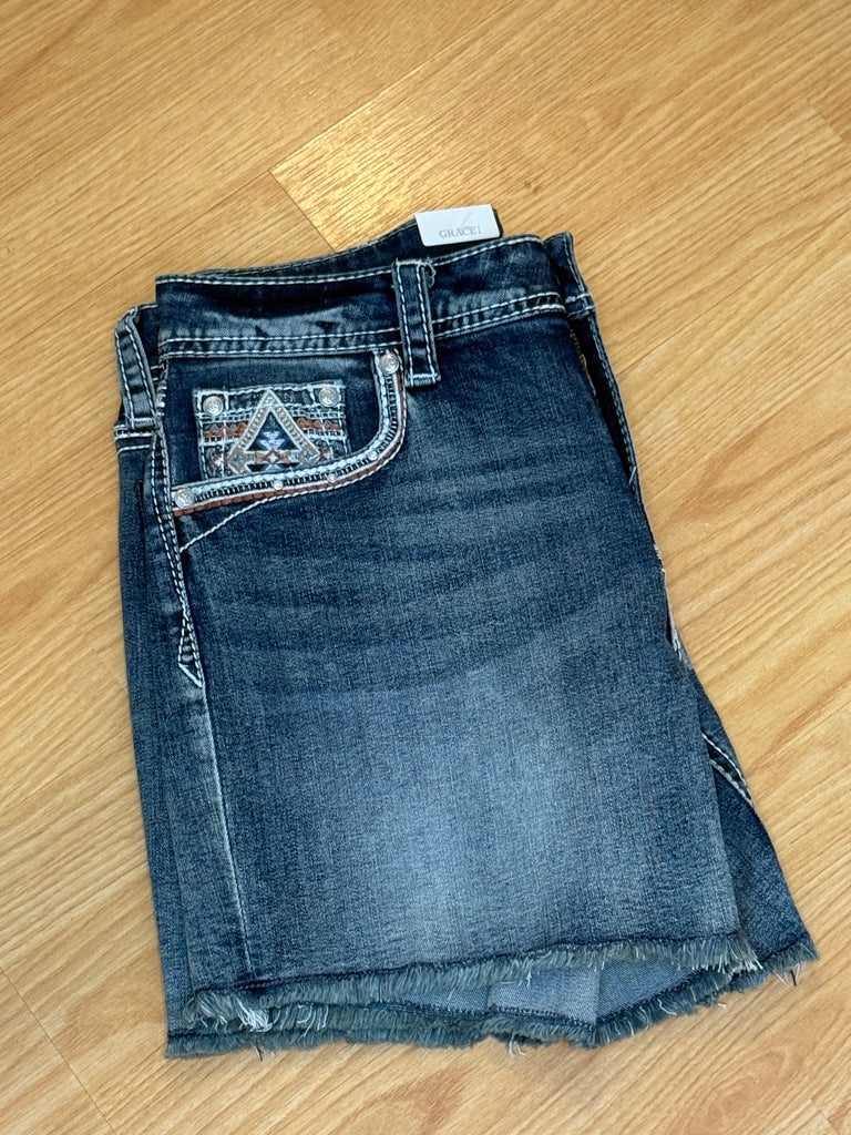A pair of denim shorts with Aztec-inspired embroidery on the back pockets and a mid-rise fit, laid flat on a wooden surface.