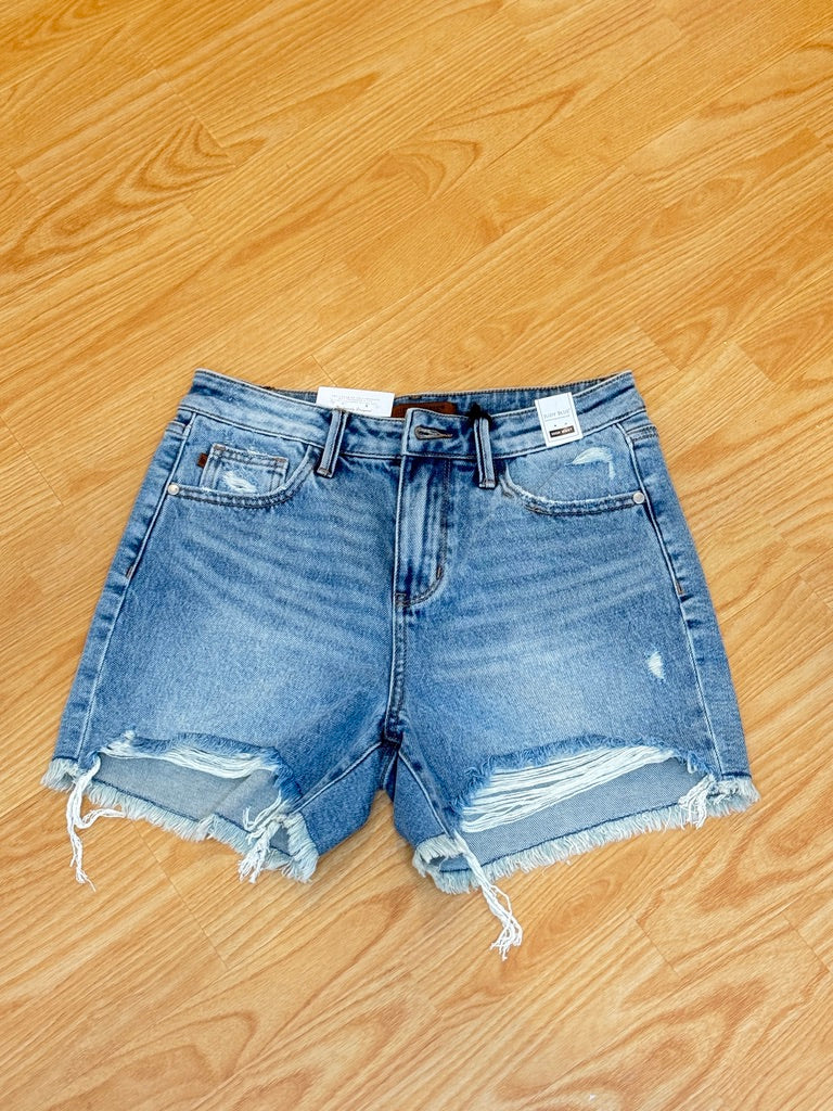 A pair of Judy Blue high-rise denim shorts with a distressed hem and pockets, displayed on a wooden surface.