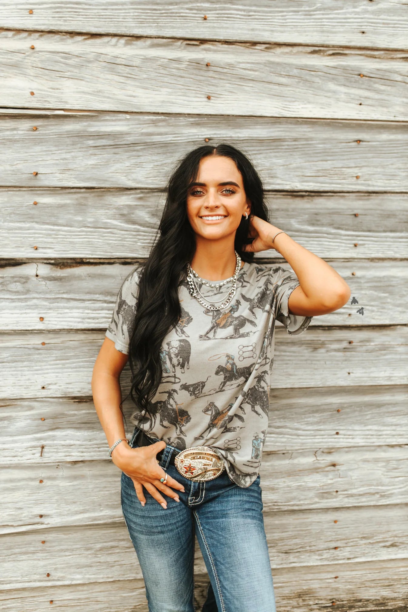 A woman smiling and posing with one hand raised to her head, wearing a short-sleeved camouflage t-shirt and blue jeans, standing in front of a wooden backdrop.
