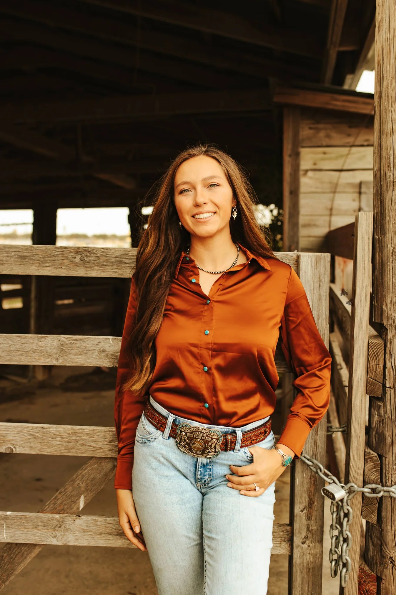 A woman wearing a saddle brown satin button-up shirt with turquoise snaps, paired with light blue jeans, standing in front of a wooden fence.