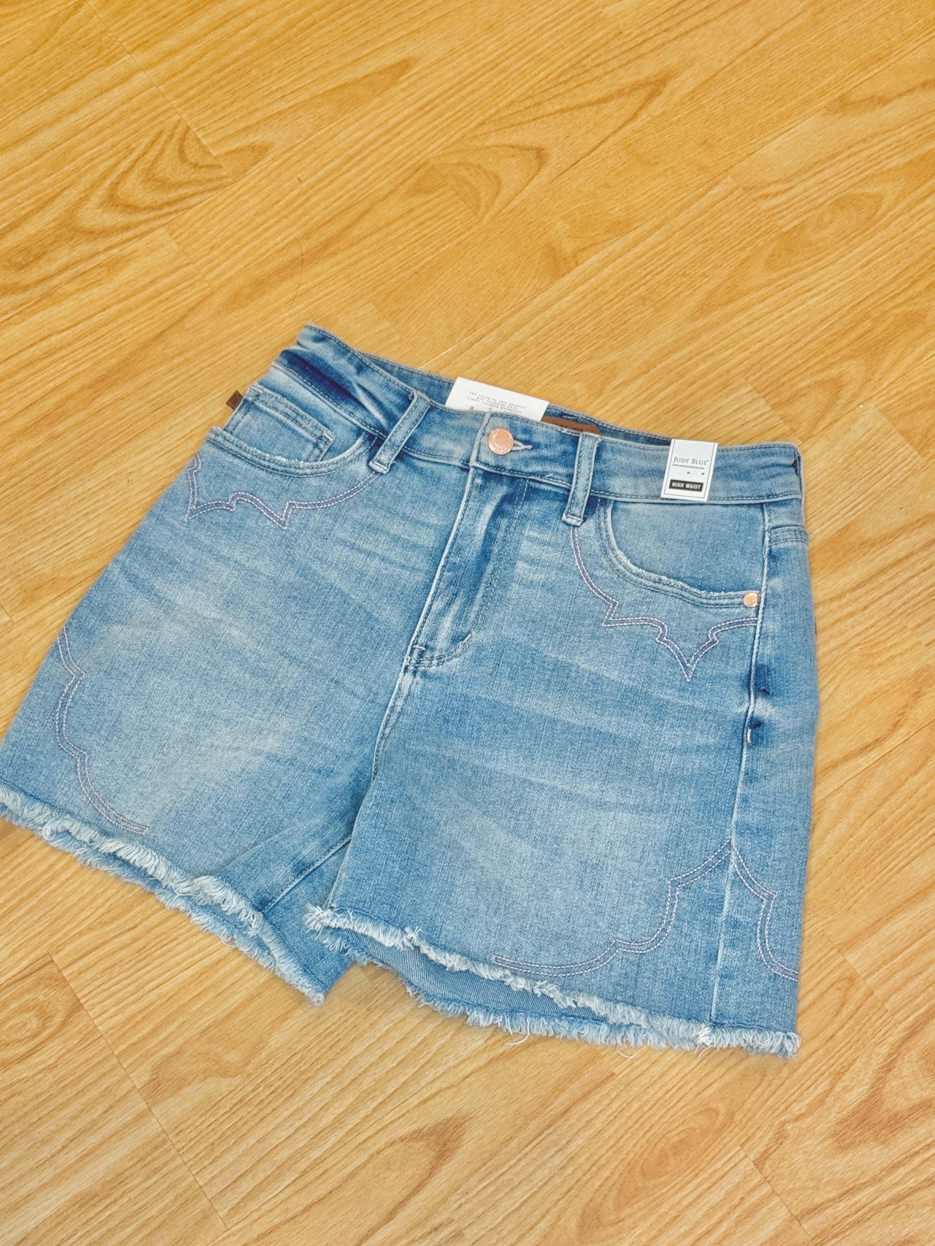 A pair of denim shorts with a mid-rise waist and relaxed fit, featuring pink embroidered stitching on the pockets and back pockets, displayed on a wooden surface.