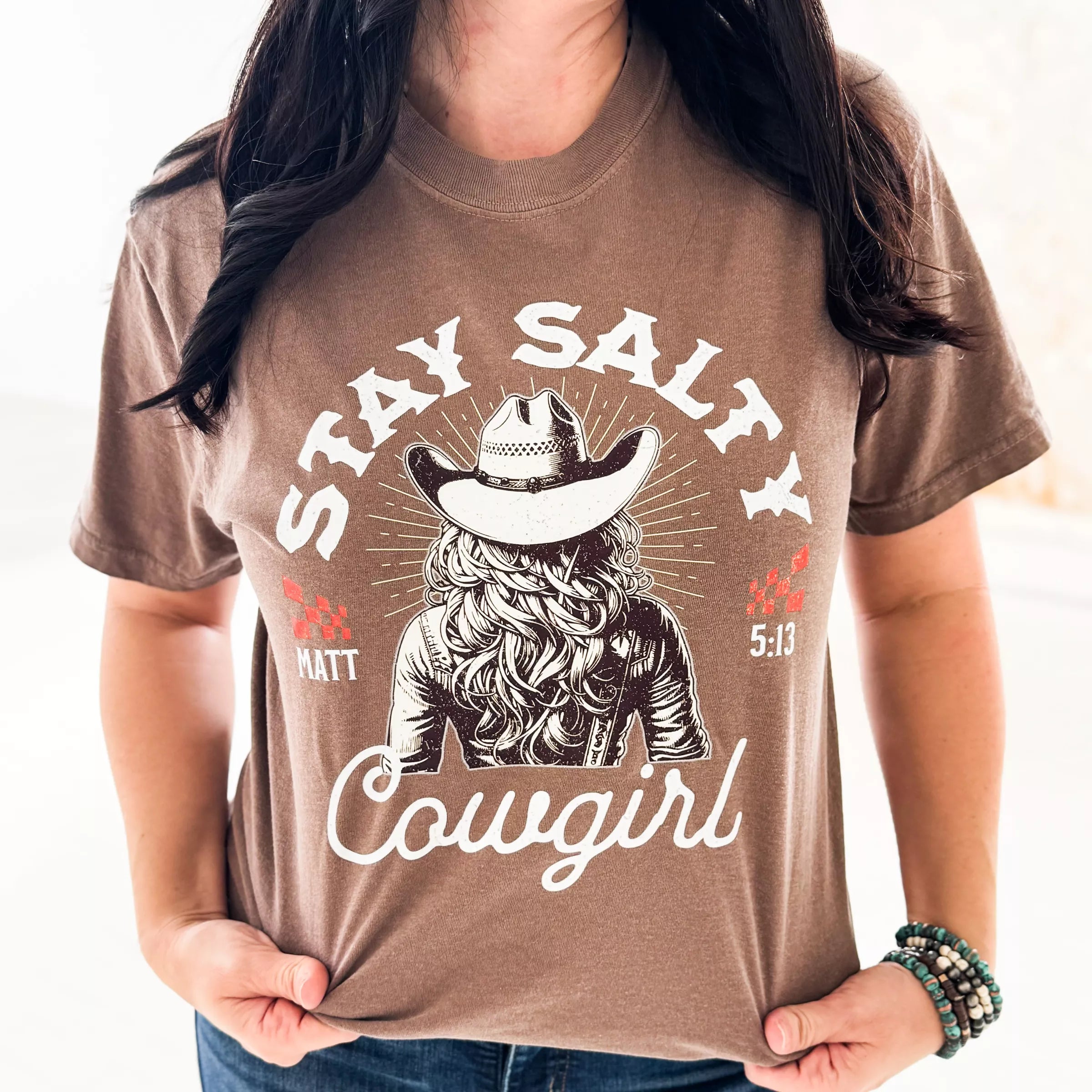 Person wearing a brown t-shirt with a graphic design and text 'Stay Salty Cowgirl' on a plain background