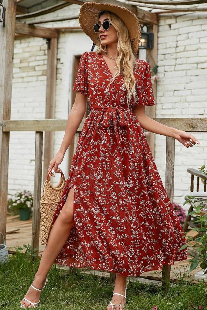 Woman in a red floral dress with a straw hat and bag, standing outdoors.