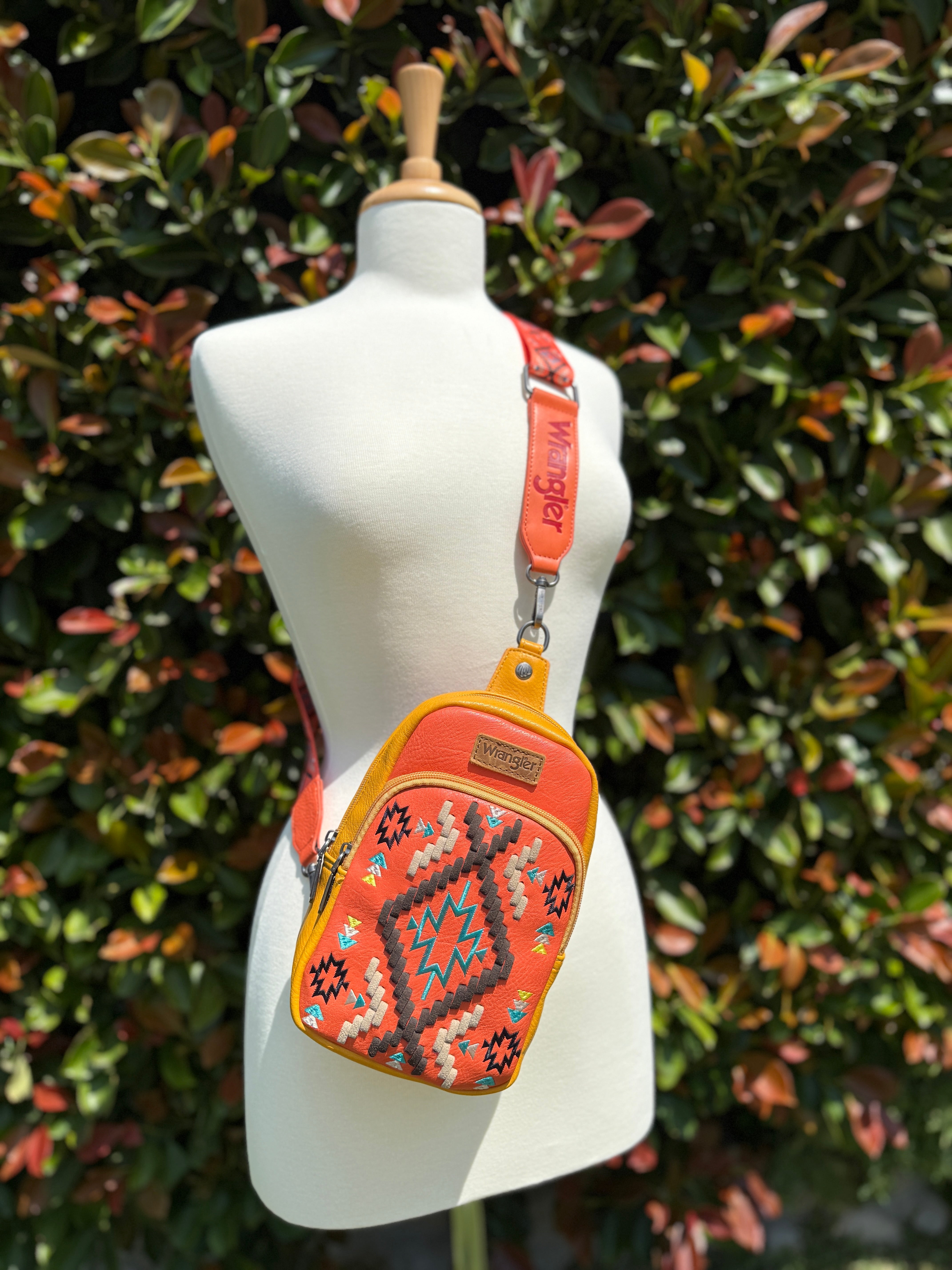 A bright orange and multicolor geometric embroidered Wrangler Southwestern sling bag displayed on a mannequin against a leafy background.