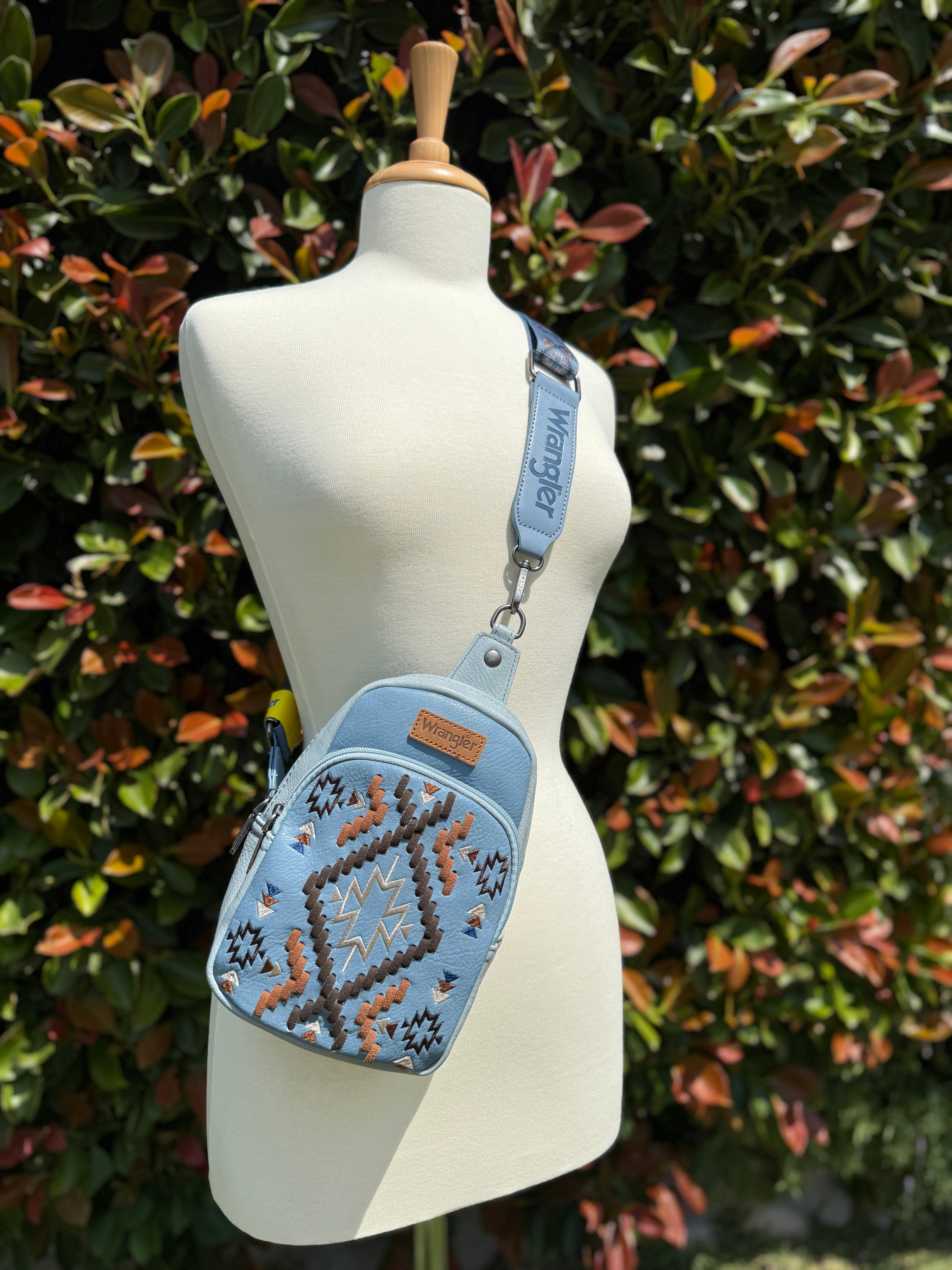A blue Wrangler Aztec Embroidered Sling Bag displayed on a mannequin against a foliage background.