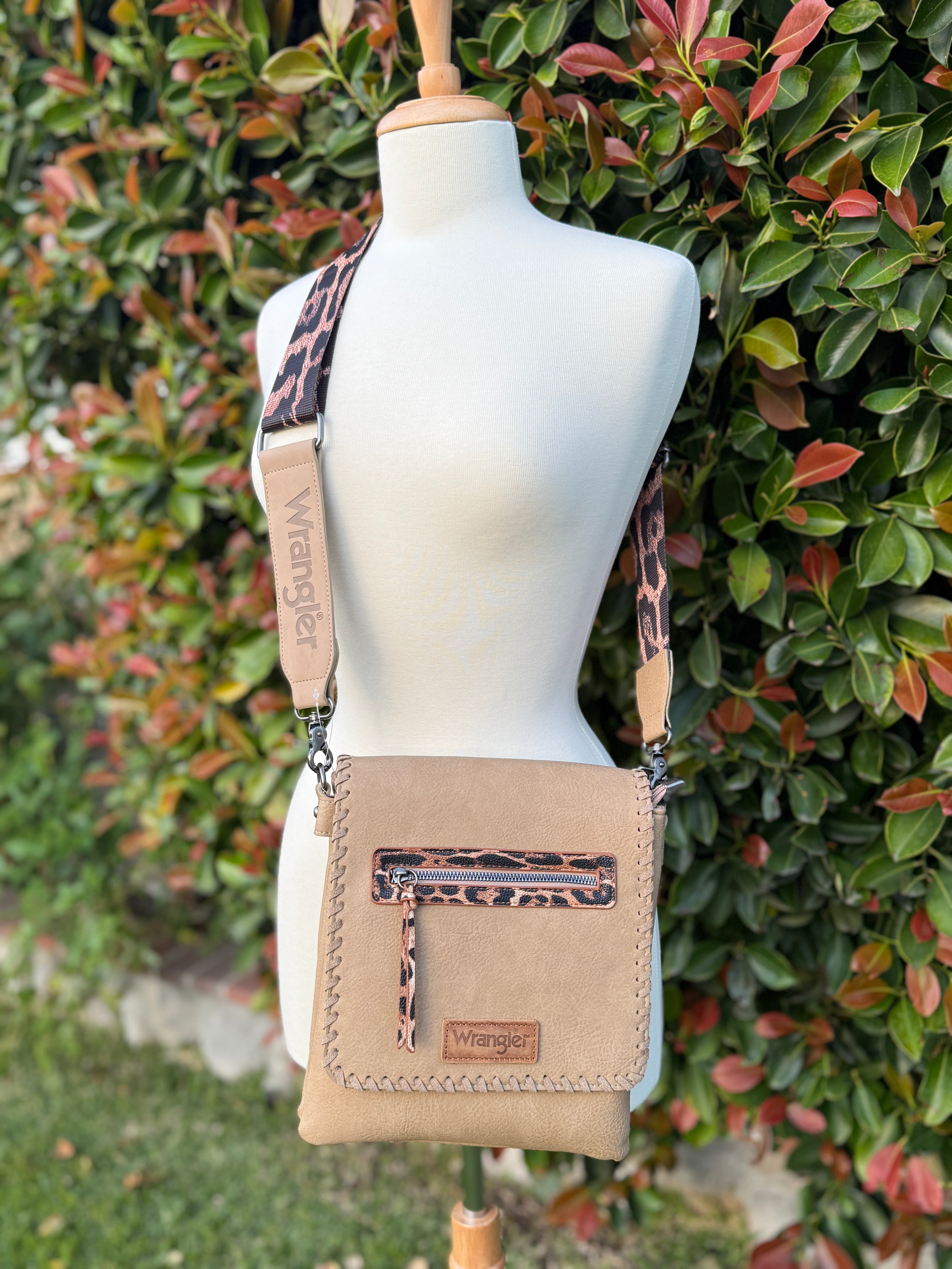 A khaki crossbody bag with whipstitch detailing and a leopard print strap, displayed on a mannequin against a foliage background.