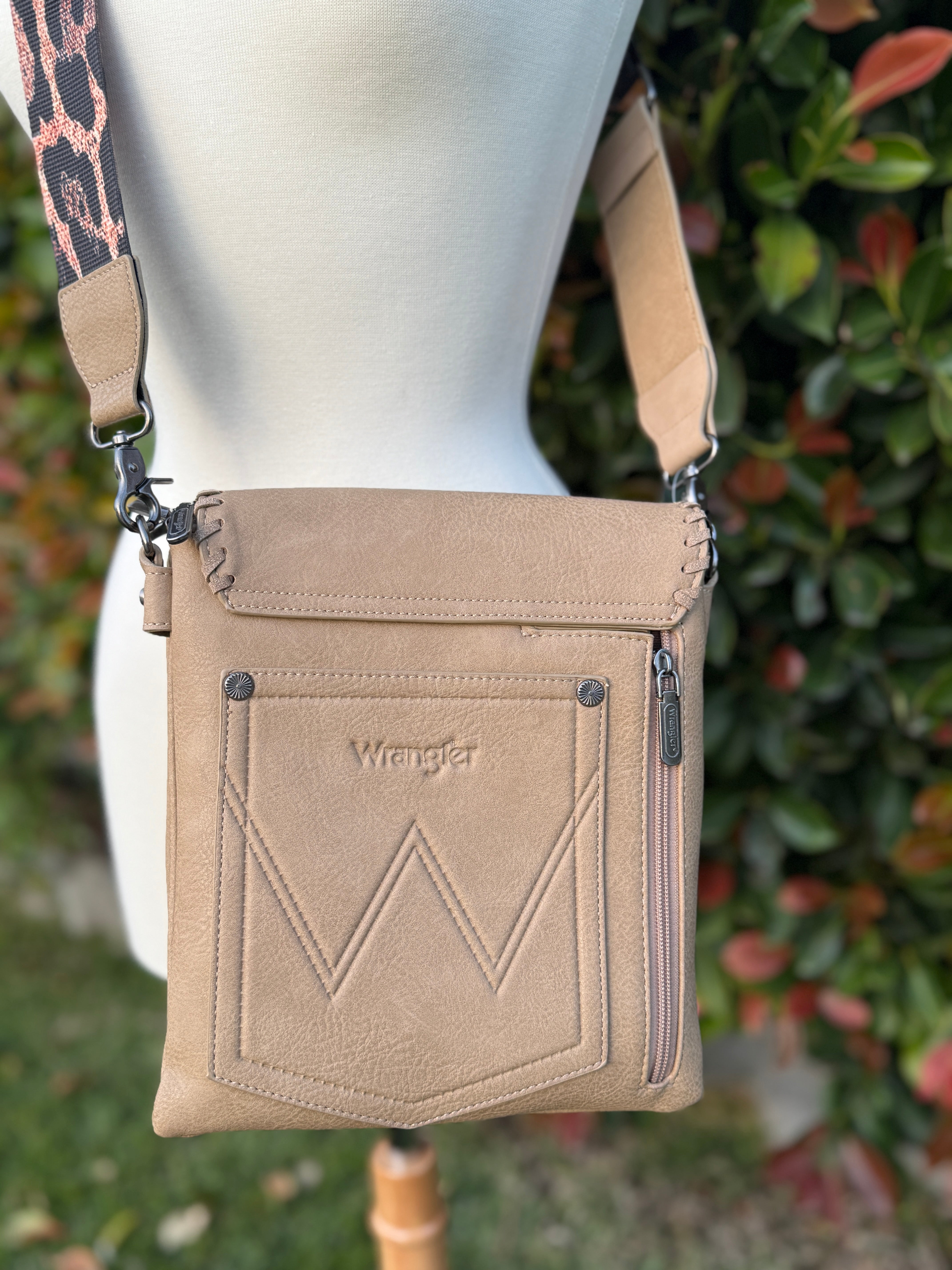 A khaki crossbody bag with whipstitch detailing and a leopard print strap, displayed on a mannequin against a foliage background.
