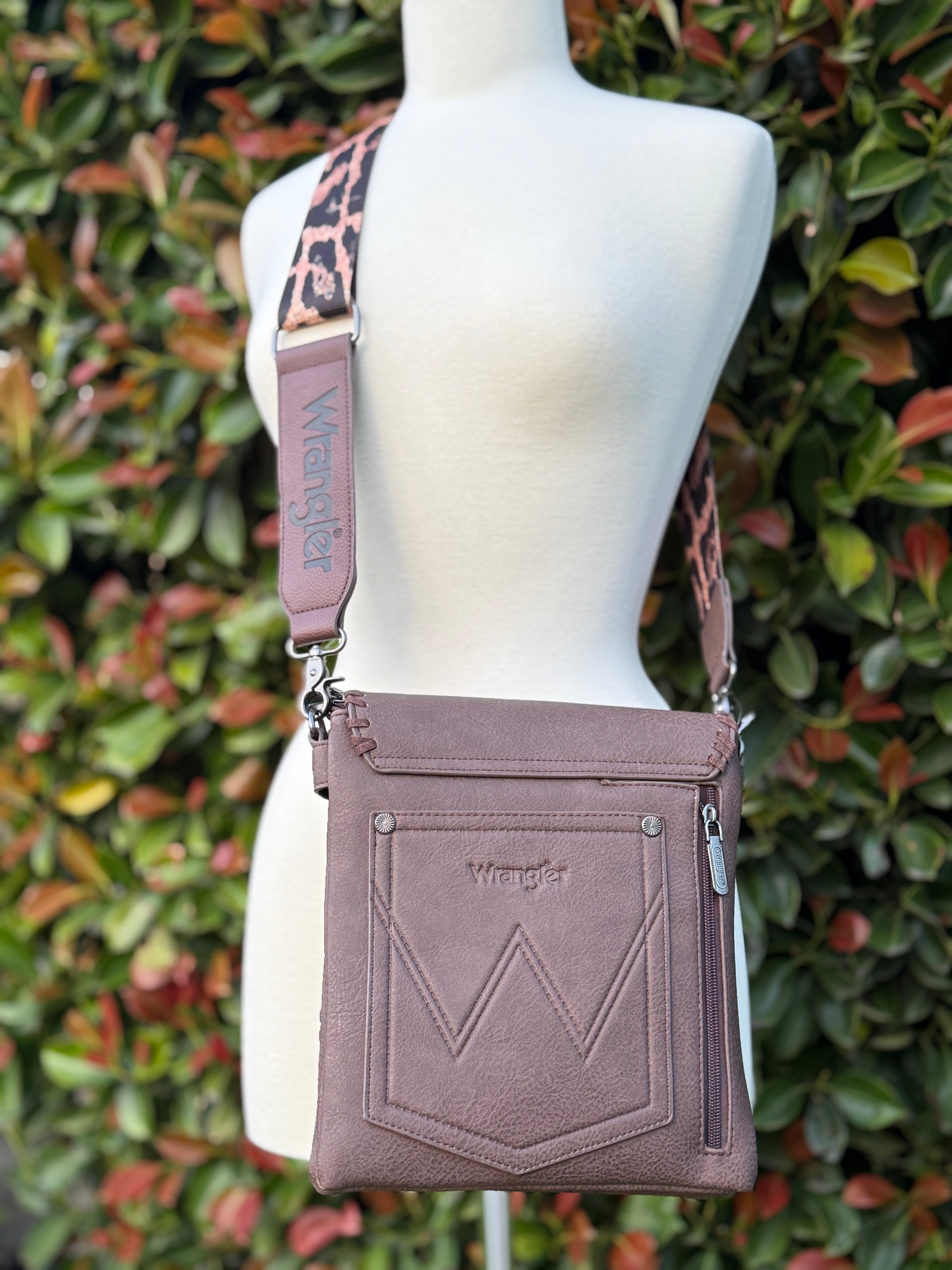 A brown crossbody bag with leopard print strap displayed on a mannequin against a leafy background.