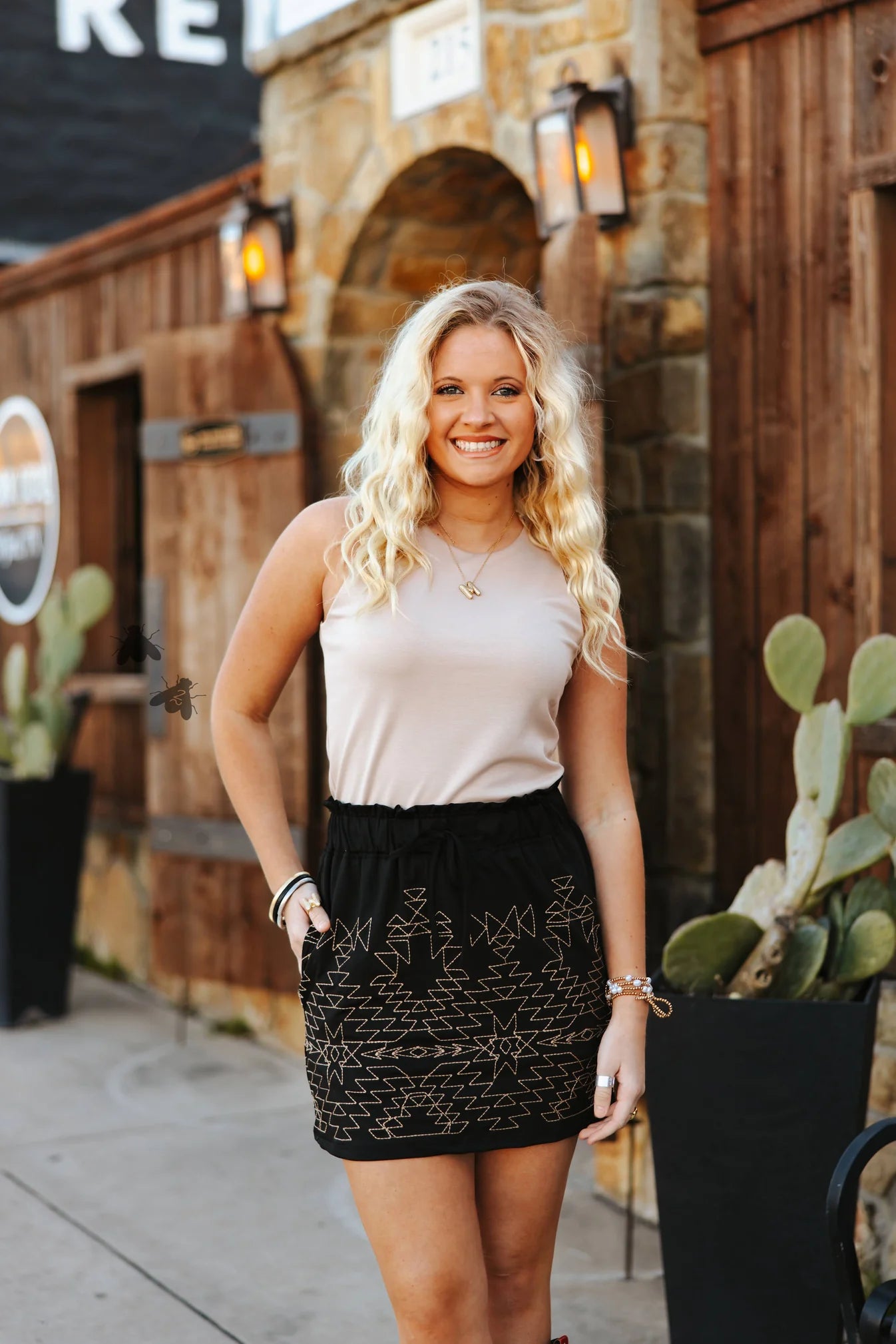 A woman standing outdoors wearing a beige sleeveless top and a black skort with a geometric pattern. She has blonde hair and is smiling at the camera.