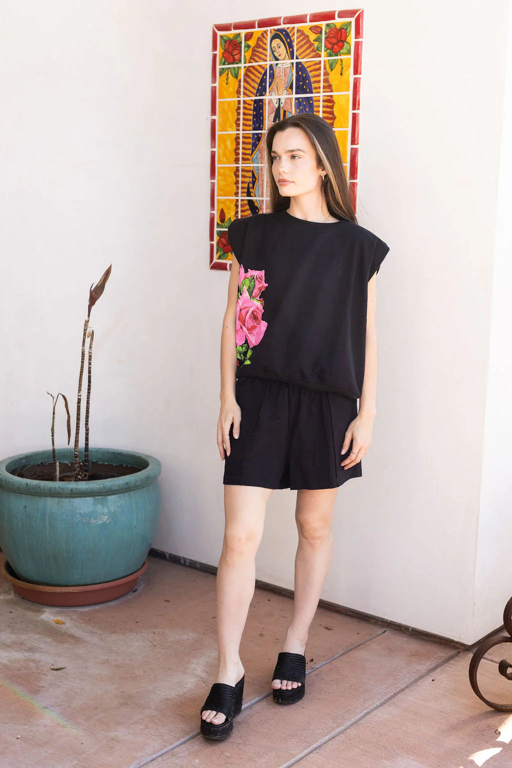 Woman wearing a black outfit with floral design in a room with a plant and colorful artwork on the wall.