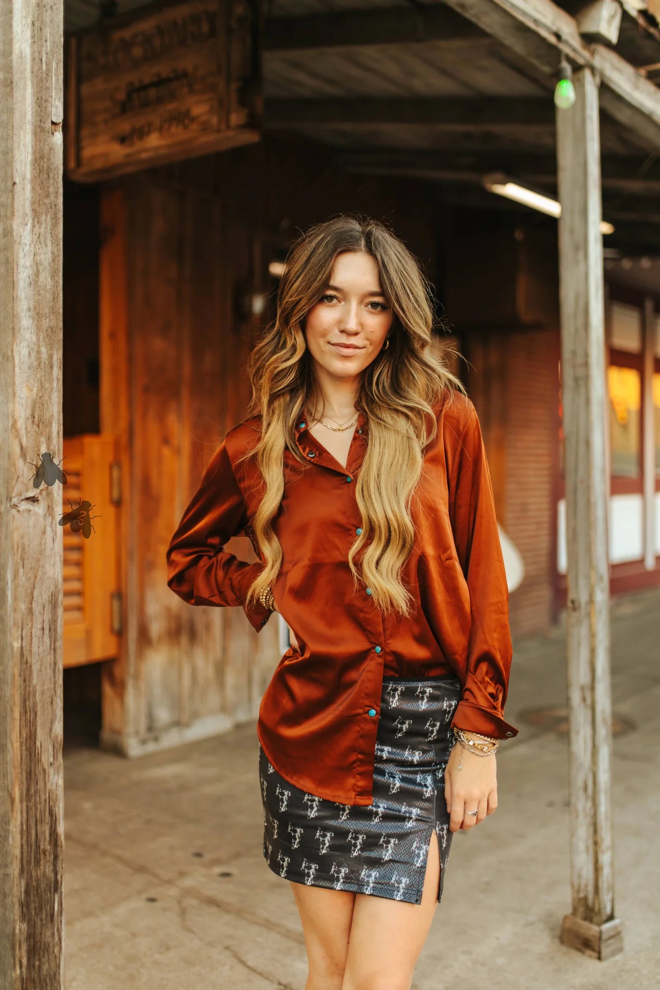 Woman wearing a rust-colored blouse and patterned skirt standing in front of a wooden building.
