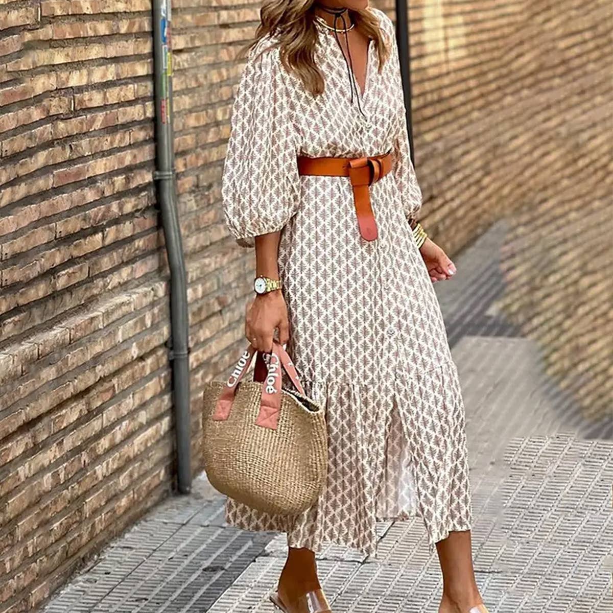 Woman in a patterned dress with a brown belt and straw bag against a brick wall.
