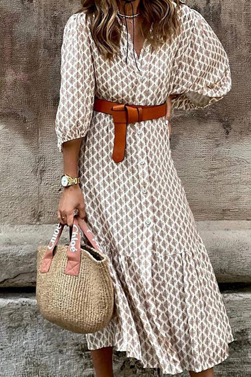 Woman wearing a patterned dress with a brown belt and holding a straw bag against a stone wall.