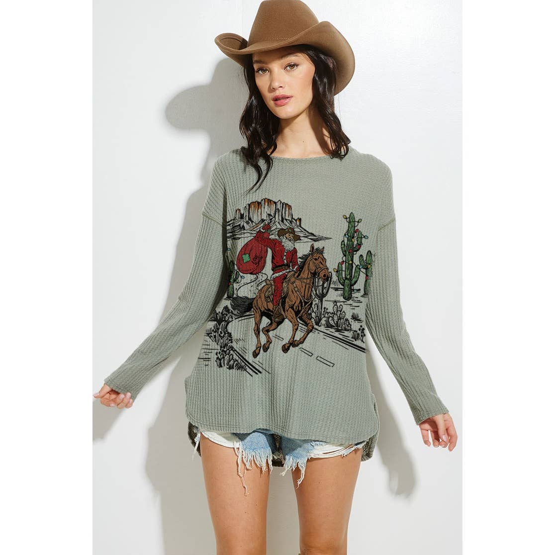 Woman wearing a green sweater with a graphic design, brown cowboy hat, and denim shorts on a white background