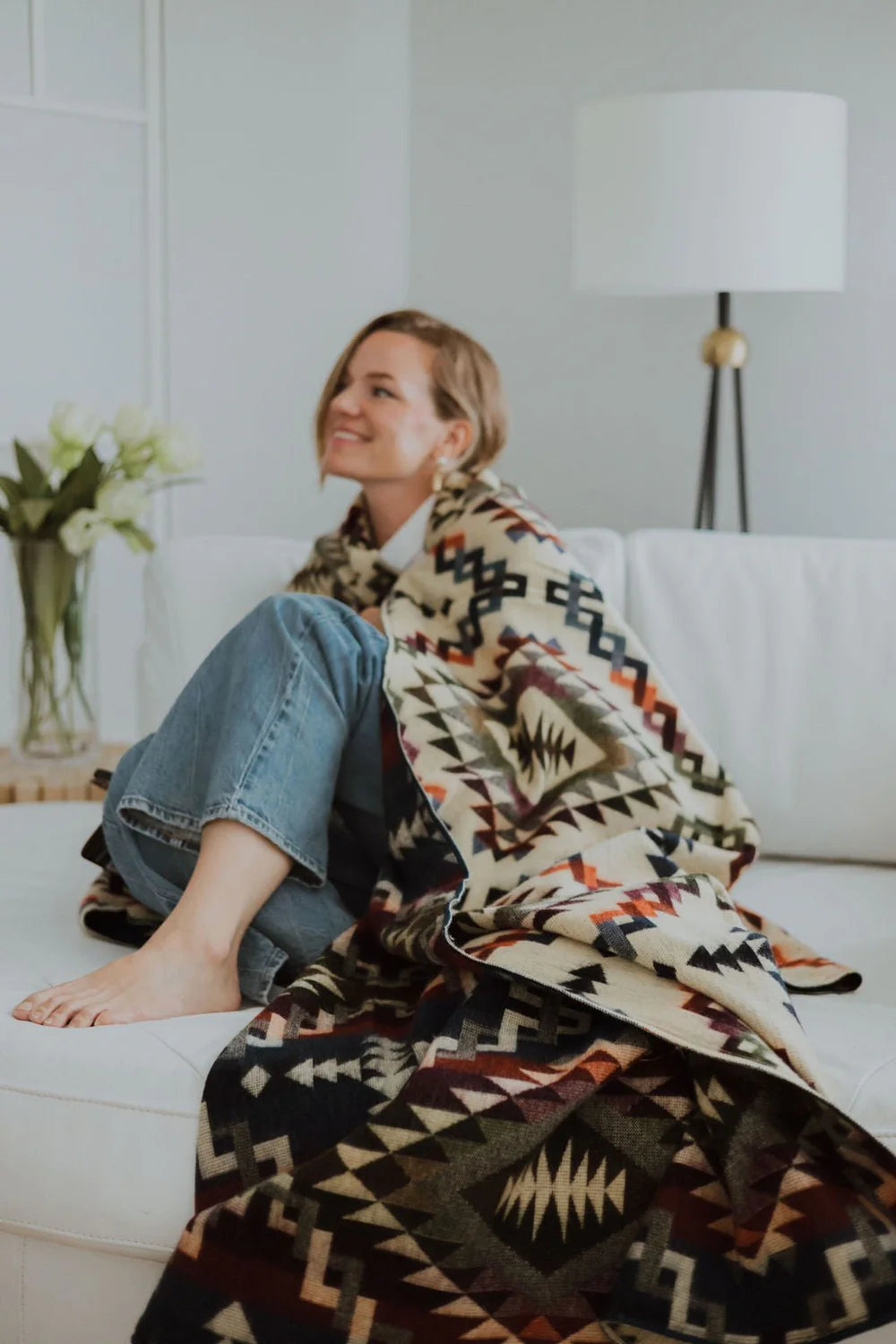 Person sitting on a couch with a patterned blanket draped over them, in a room with a lamp and flowers.