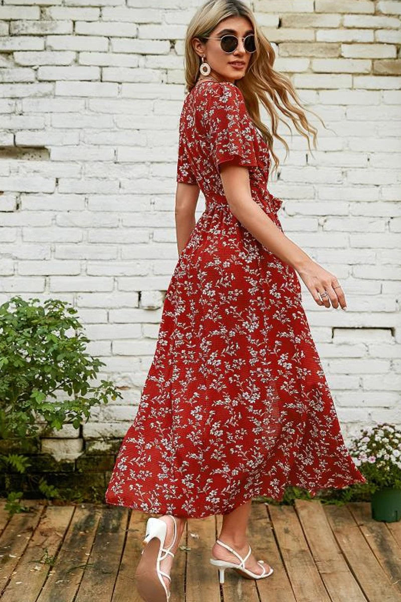 Woman wearing a red floral dress standing against a white brick wall.