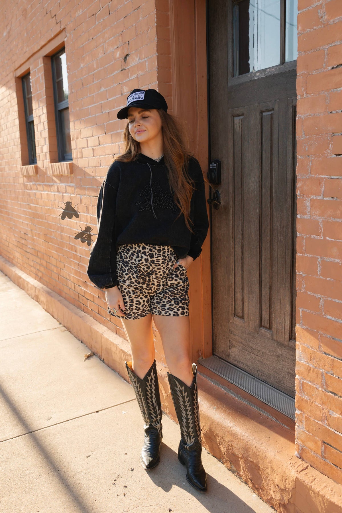 Woman in black top, leopard print shorts, and cowboy boots standing in front of a brick building.