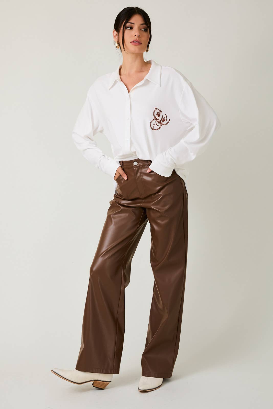 Woman wearing a white blouse with a small design and brown leather pants on a plain background
