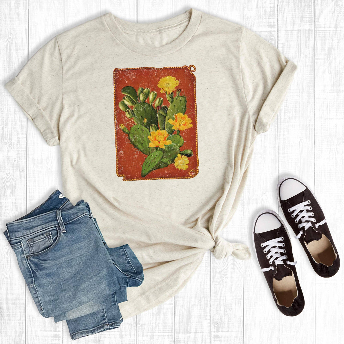 A styled flat-lay of a light beige graphic T-shirt with rolled sleeves, featuring a vintage-style red and green cactus print. The shirt is styled with a pair of blue jeans and black canvas sneakers on a white wood plank background.