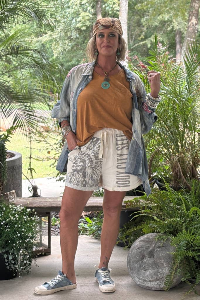 Woman standing outdoors in a garden setting wearing a mustard top, denim jacket, and patterned shorts.