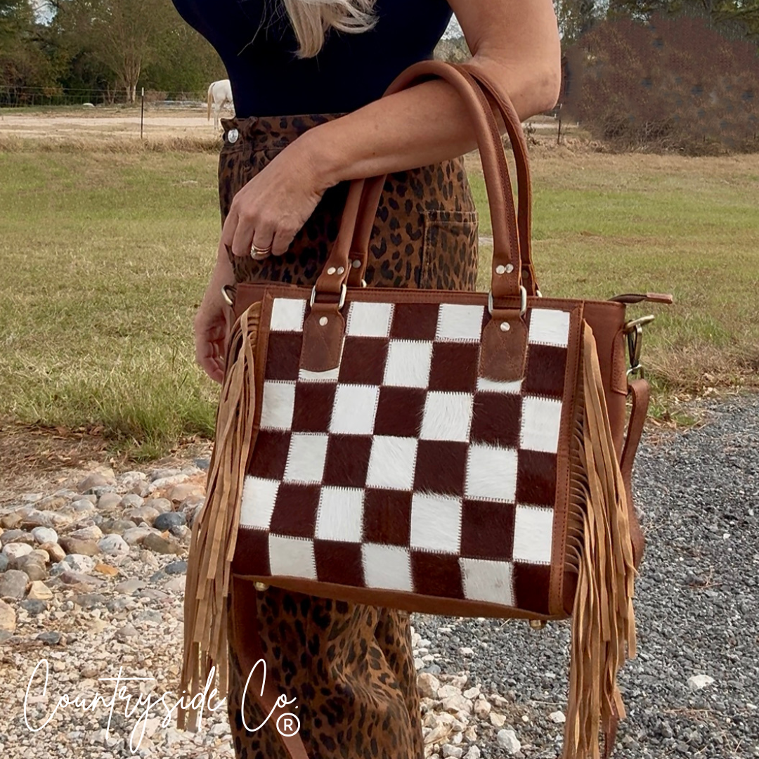 Person holding a brown and white checkered handbag with fringes outdoors.