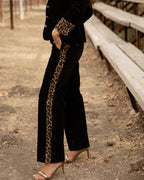 A person wearing black wide-leg pants with a tan and black leopard print stripe down the side, paired with a matching black jacket and gold heeled sandals, standing outdoors on a dirt path beside a wooden bench.