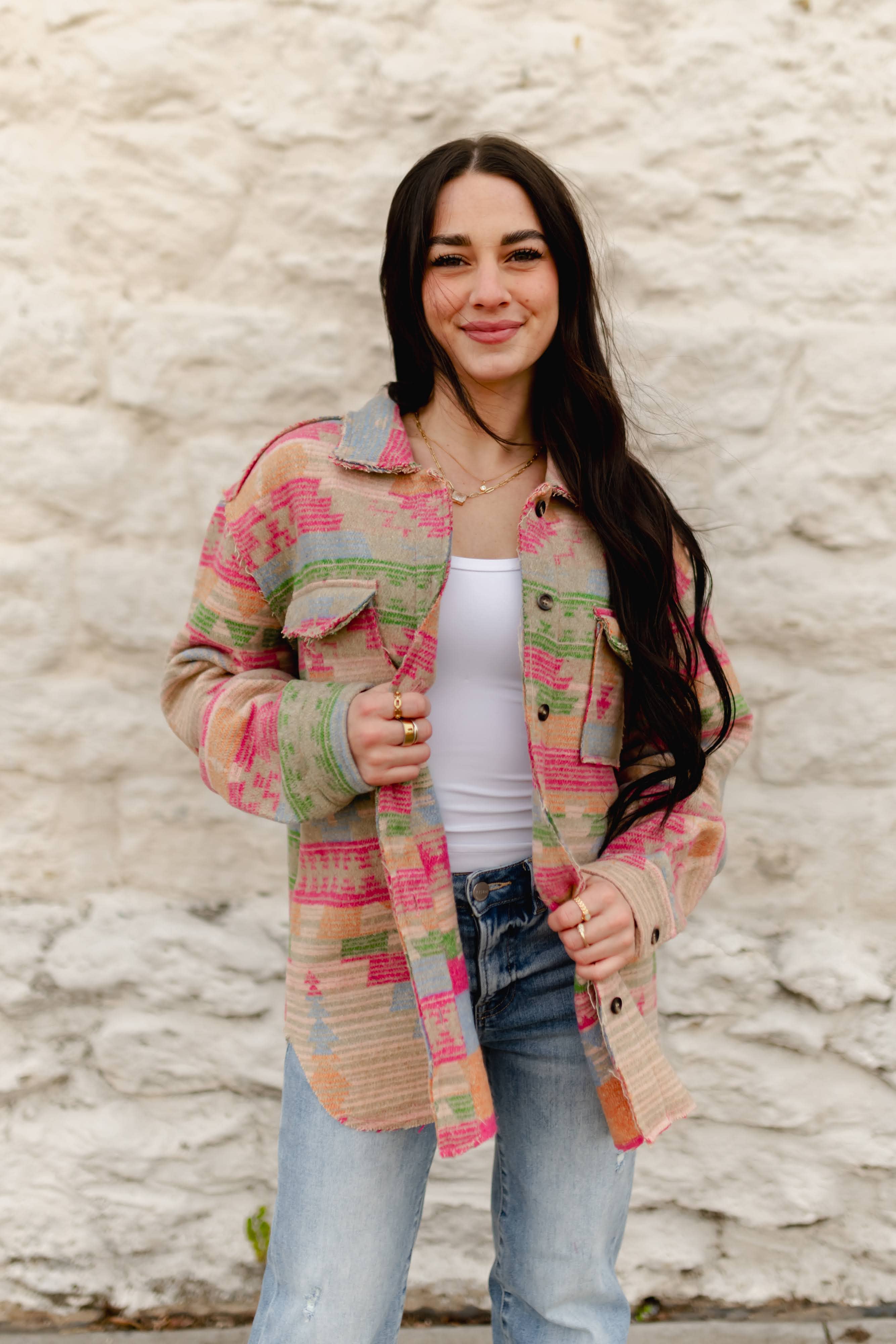 A woman with long dark hair stands against a textured white stucco wall wearing a colorful patchwork button-up shirt jacket over a white tank top and light-wash blue jeans.
