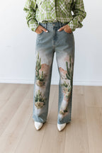 A person wearing light-wash blue jeans with a desert landscape print featuring cacti and pink mountains, styled with a green plaid shirt and white pointed shoes.