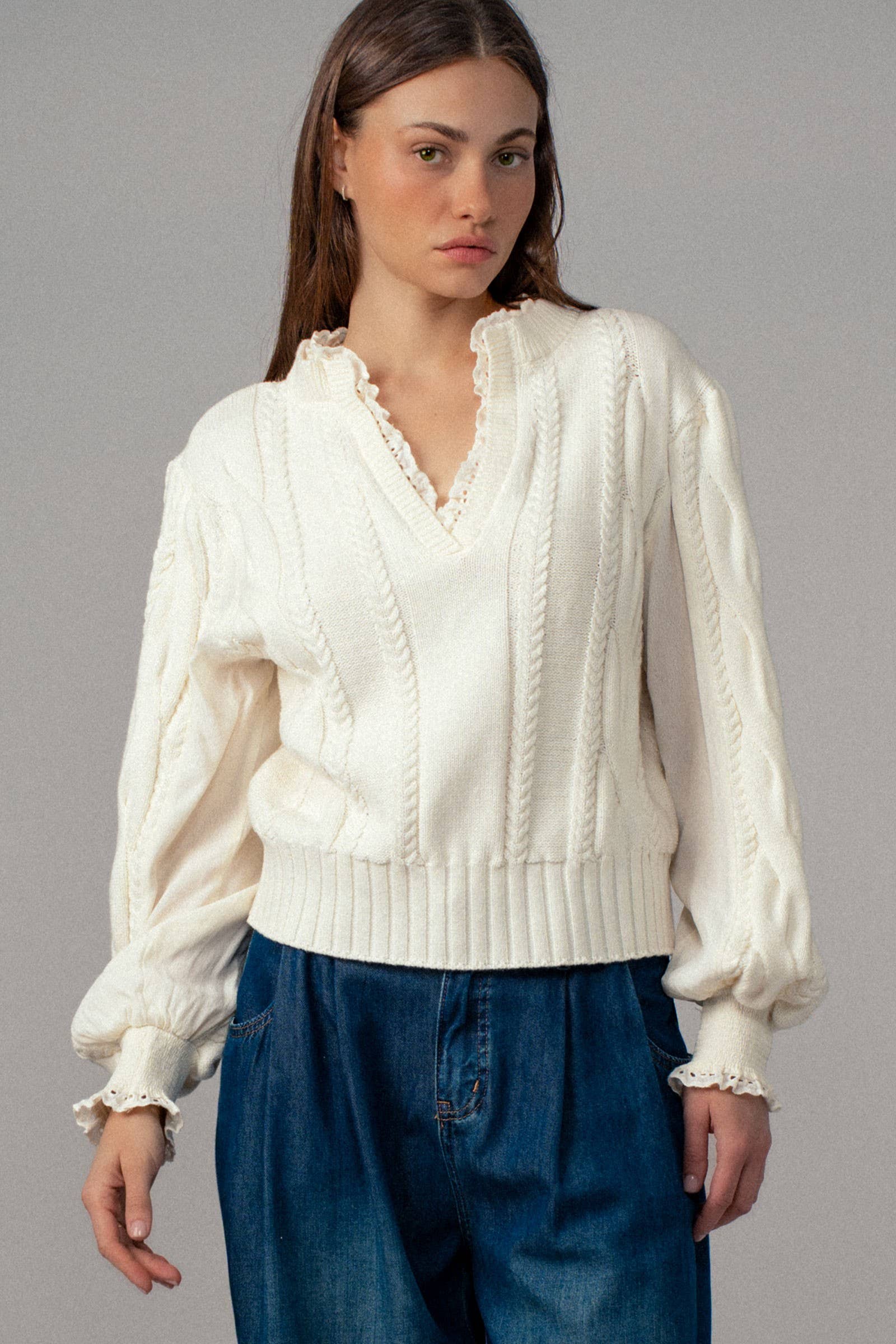 Woman wearing a white cable knit sweater and blue jeans against a gray background