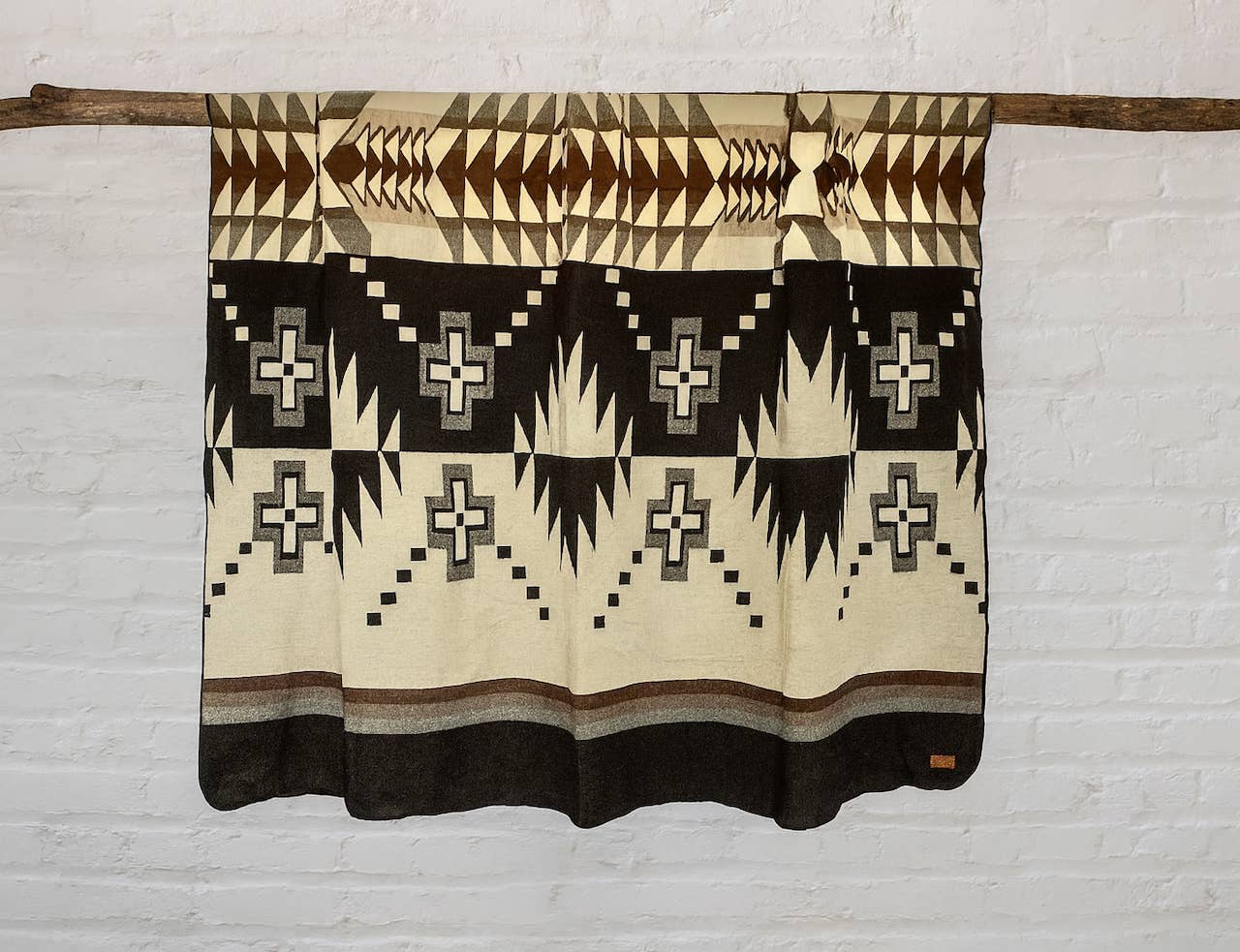 A woven southwestern-style blanket with geometric patterns in black, cream, and brown, hanging from a wooden dowel against a white brick wall.