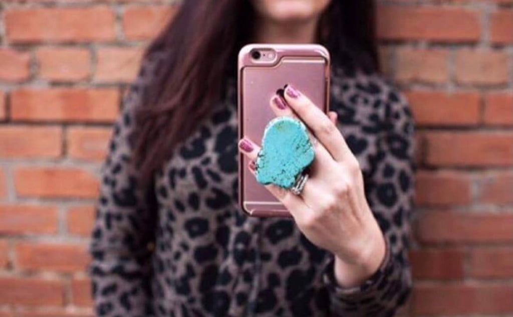 A person holding a rose gold smartphone with a textured turquoise pop socket, standing in front of a red brick wall.