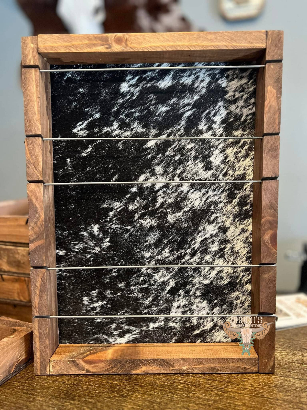 A rustic wooden horse stall or tack box with a black and white speckled hide panel and five horizontal metal wires, shown on a wooden table.