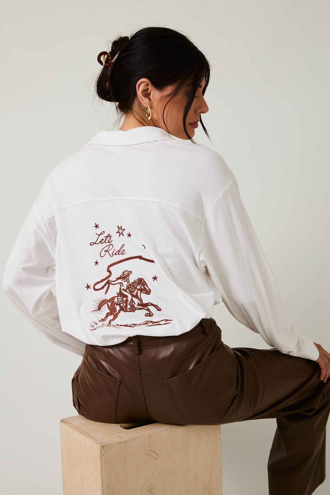 Person wearing a white shirt with embroidery sitting on a wooden block against a plain background