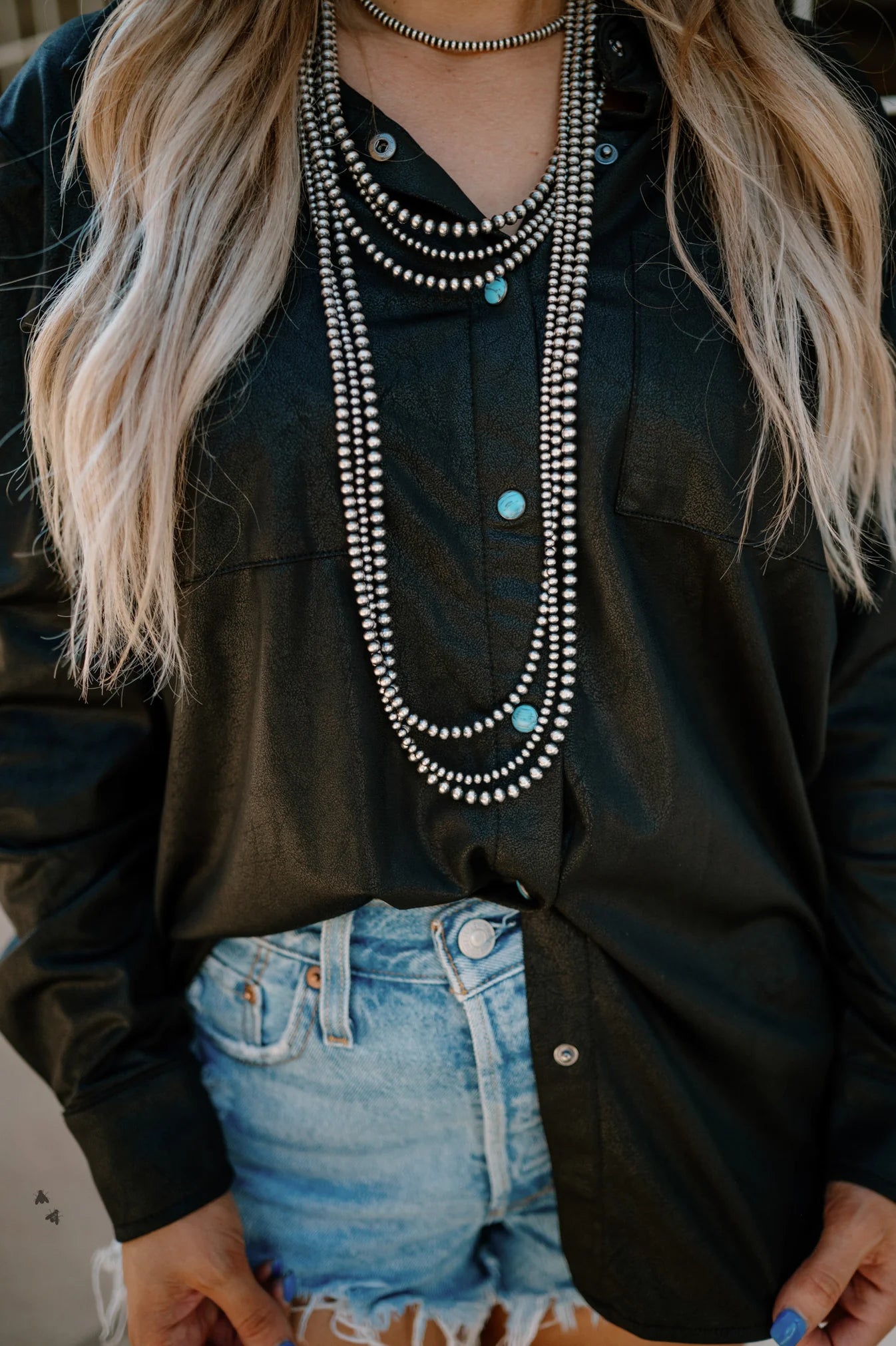 A person wearing a black leather button-down shirt over a white top, paired with blue denim shorts, accessorized with multiple necklaces including a turquoise pearl snap necklace.