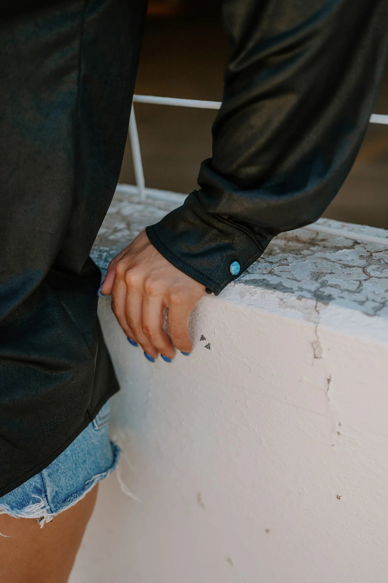 Person wearing a dark jacket and denim shorts leaning on a wall.