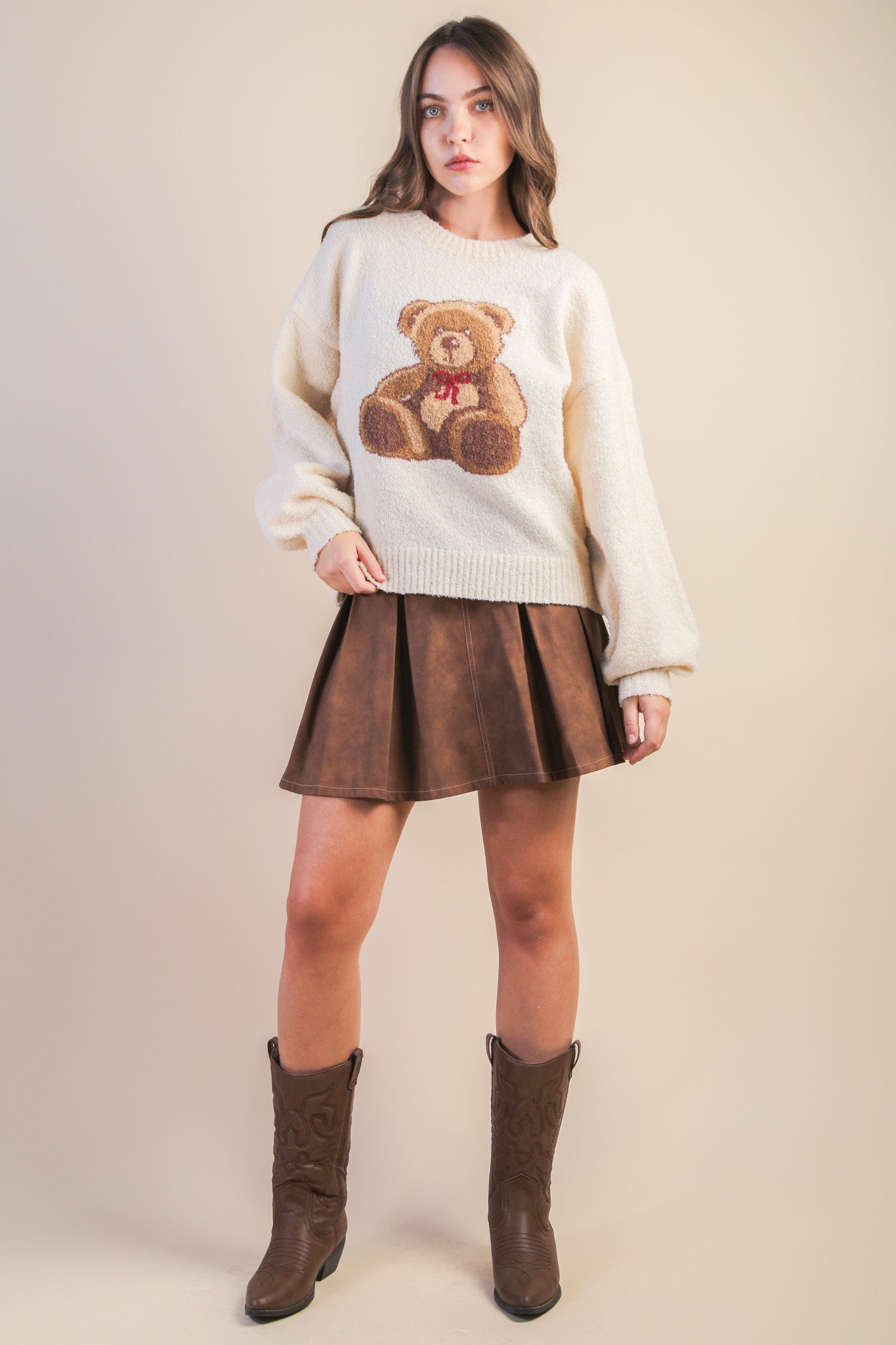 Woman wearing a cream sweater with a teddy bear design, brown pleated skirt, and brown boots on a beige background