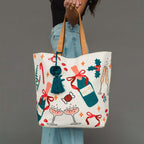 A person holds a large cream canvas tote bag decorated with Christmas-themed illustrations including champagne bottles, champagne glasses, candy canes, holly, and red bows. The bag has tan shoulder straps and a dark teal tassel charm.