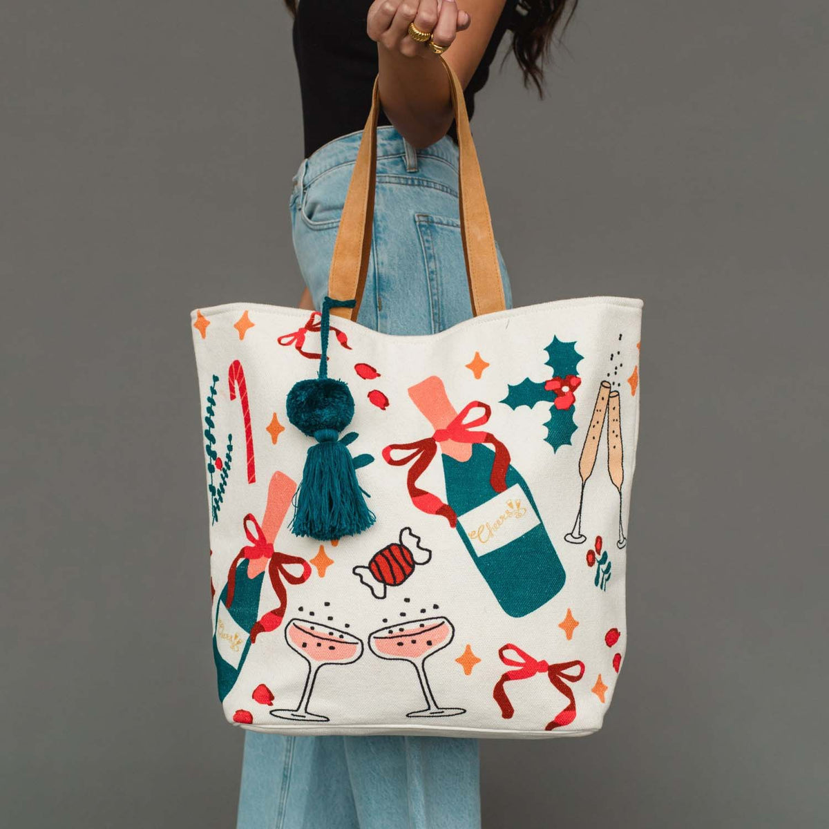 A person holds a large cream canvas tote bag decorated with Christmas-themed illustrations including champagne bottles, champagne glasses, candy canes, holly, and red bows. The bag has tan shoulder straps and a dark teal tassel charm.