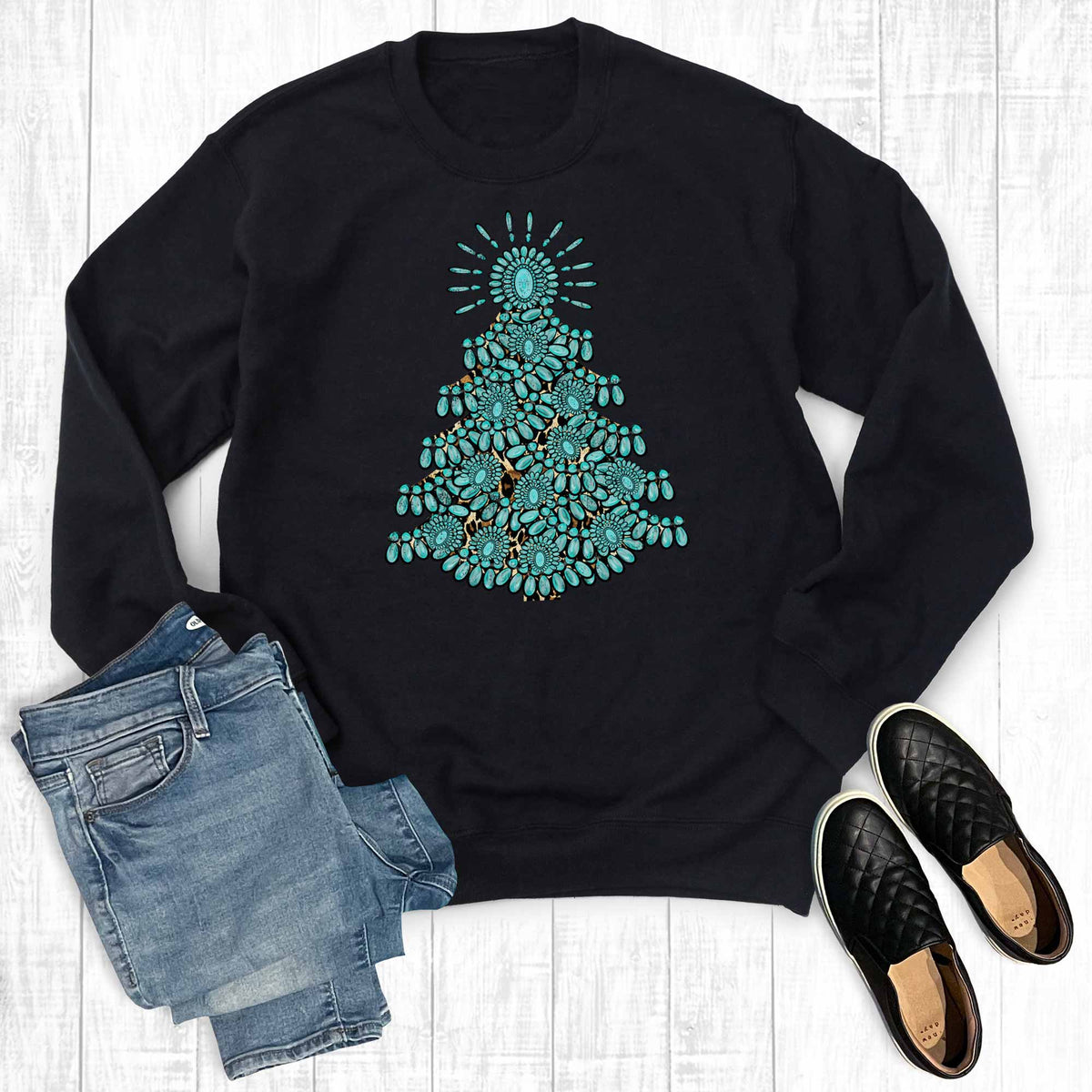 Black sweatshirt with a blue Christmas tree design, paired with denim shorts and black shoes on a white wooden surface.