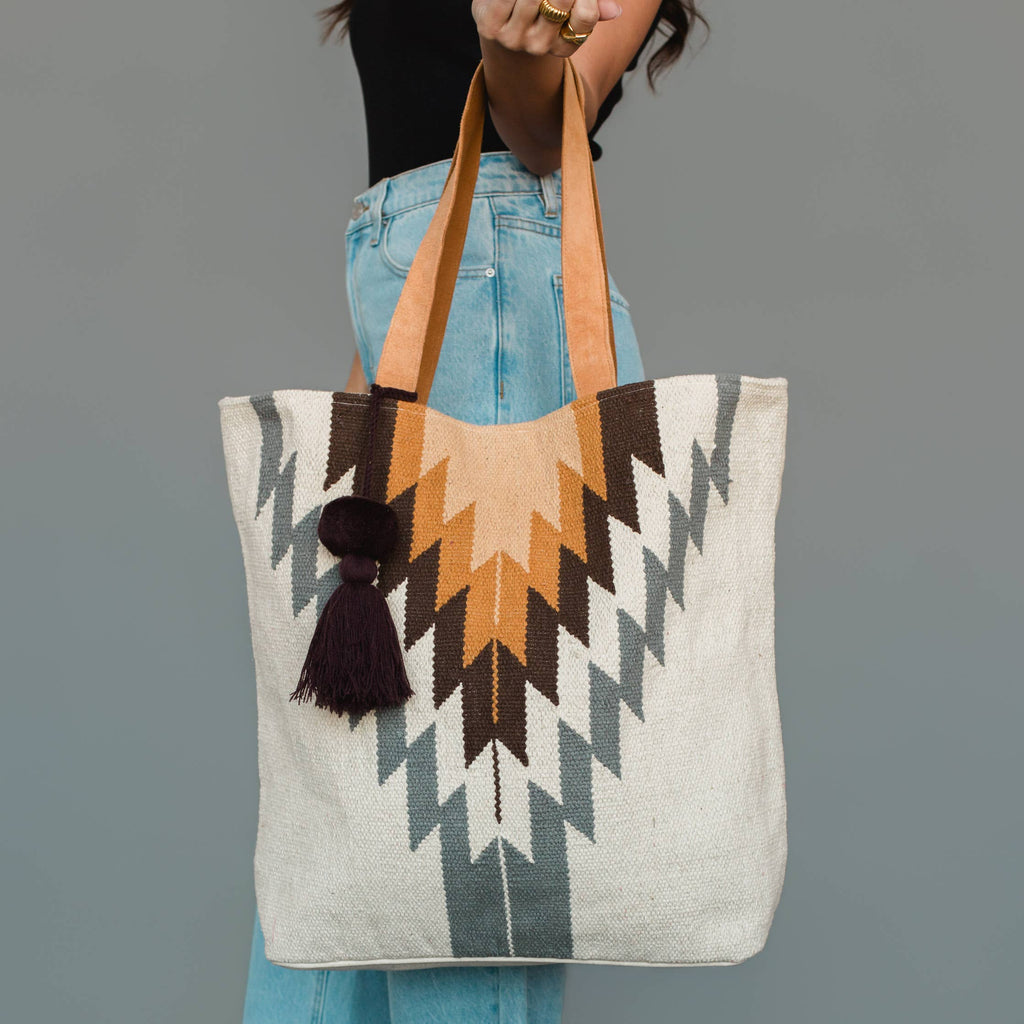 A person holds a large woven tote bag with a geometric Southwestern-style pattern in beige, brown, gray, and black, featuring tan shoulder straps and a dark tassel, against a solid gray background.