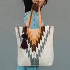 A person holds a large woven tote bag with a geometric Southwestern-style pattern in beige, brown, gray, and black, featuring tan shoulder straps and a dark tassel, against a solid gray background.