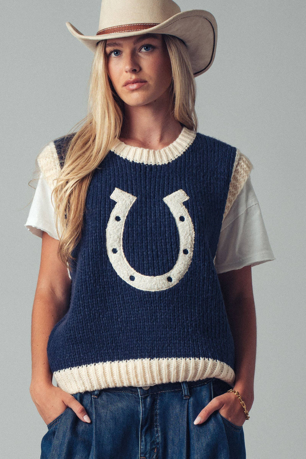 Woman wearing a navy blue sleeveless sweater with a horseshoe design, white shirt, and cowboy hat on a gray background.