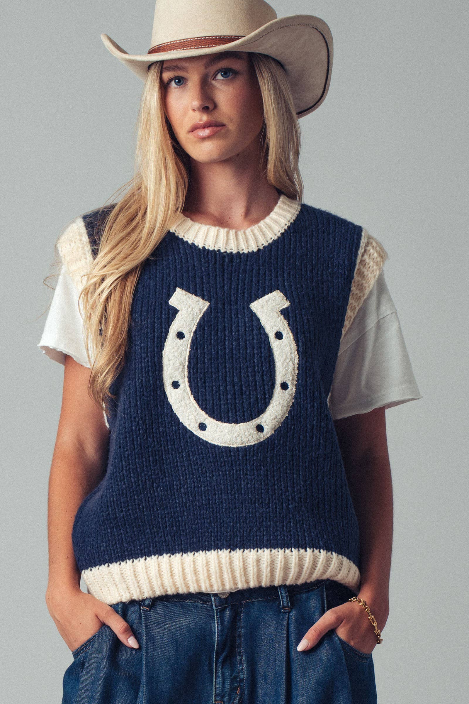 Woman wearing a navy blue sleeveless sweater with a horseshoe design, white shirt, and cowboy hat on a gray background.
