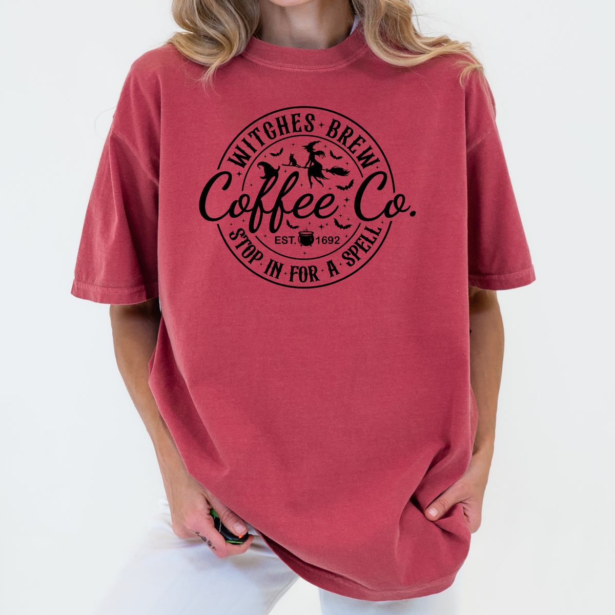 A woman wearing an oversized dusty rose red t-shirt with a black circular graphic that reads 'Witches Brew Coffee Co. Stop In For A Spell'.