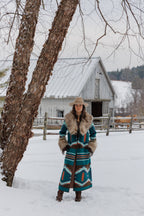 A woman stands in a snowy outdoor setting wearing a long Southwestern-style patterned coat with a fur collar and a cowboy hat, with a rustic white barn and tree in the background.