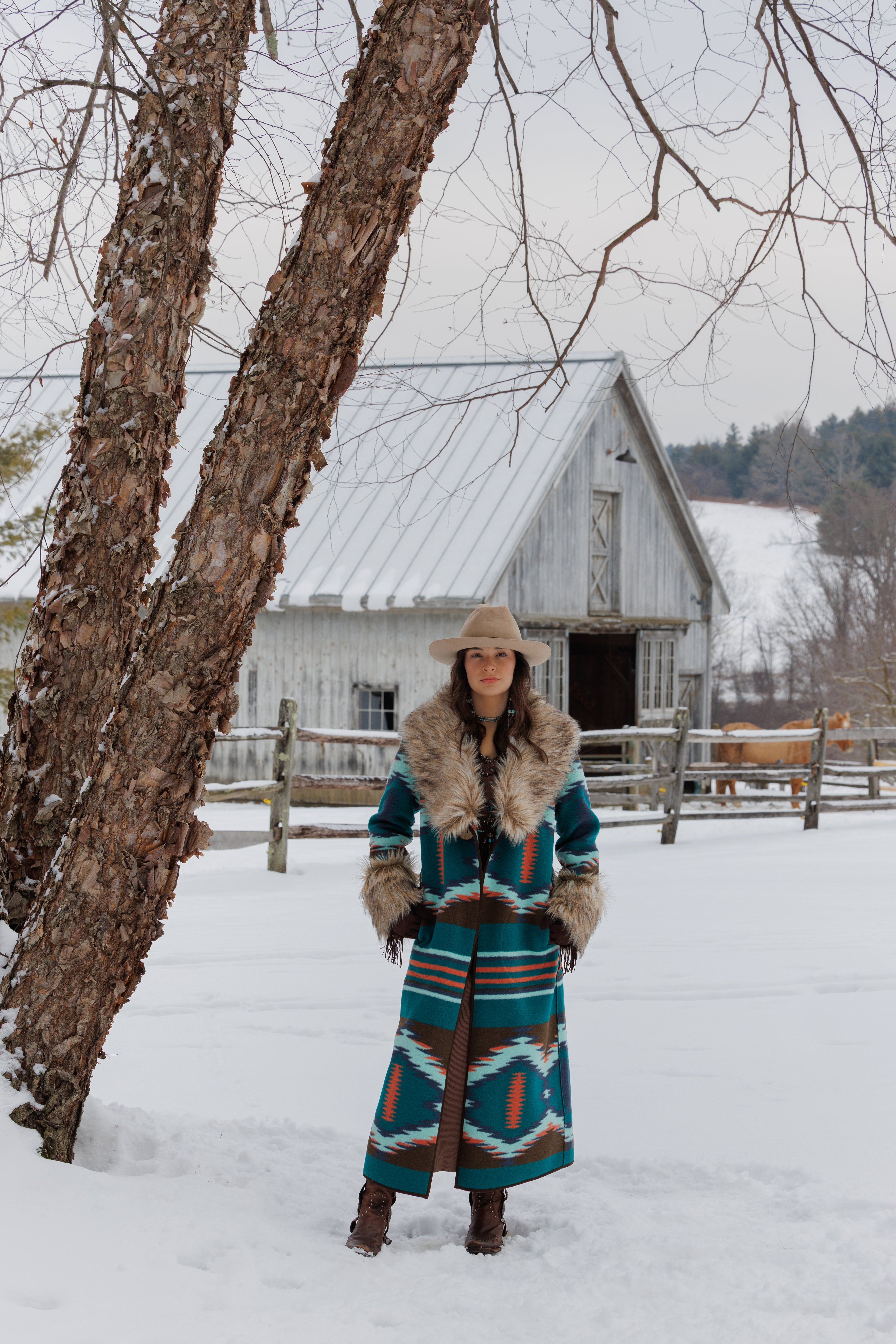 A woman stands in a snowy outdoor setting wearing a long Southwestern-style patterned coat with a fur collar and a cowboy hat, with a rustic white barn and tree in the background.
