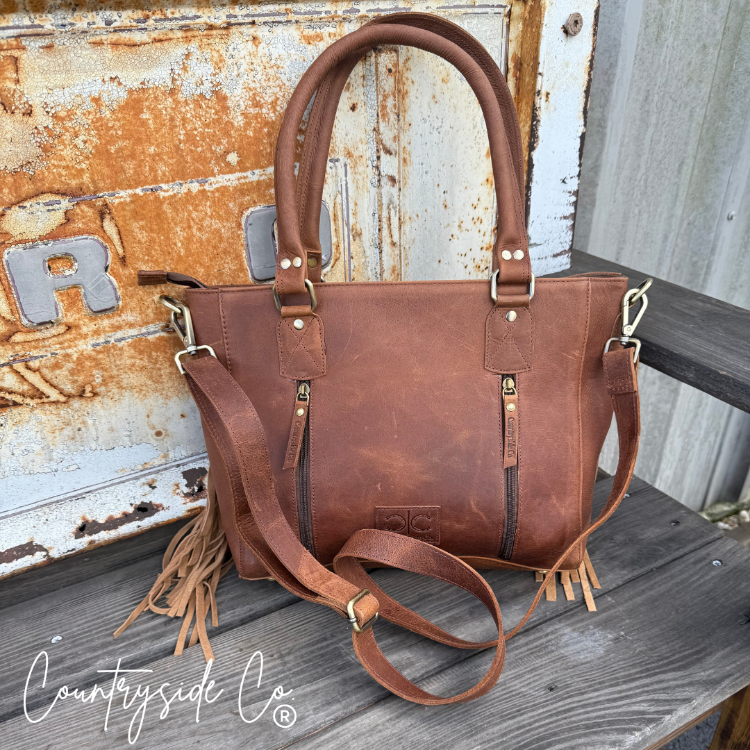 Brown leather handbag with fringe details on a rustic wooden surface, featuring the 'Countryside Co' brand.