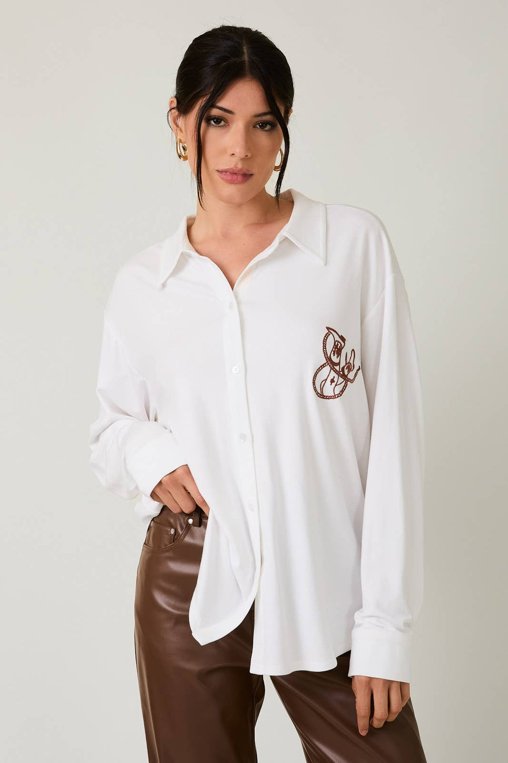 Woman wearing a white shirt with a brown emblem and brown pants on a plain background
