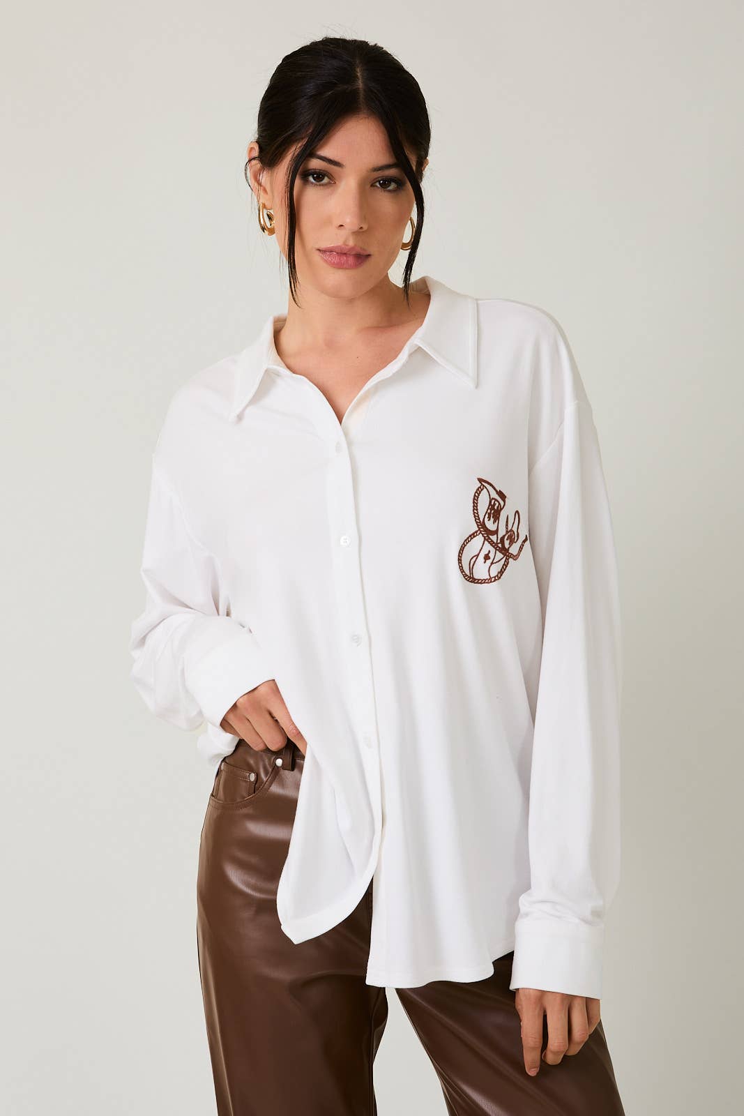 Woman wearing a white shirt with a brown emblem and brown pants on a plain background