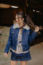 Woman wearing a denim jacket with fur trim and a graphic t-shirt, standing outdoors.